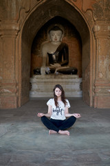 A pretty woman is meditating in lotus position, sitting on the floor in front of a Buddha statue in an ancient temple in Bagan in Myanmar.