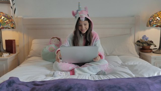 peque&ntilde;a con pijama de unicornio sentada en cama blanca realiza videoconferencia en port&aacute;til gris