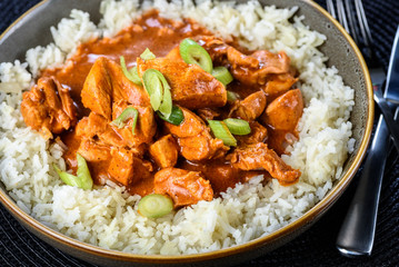 A serve of traditional Butter Chicken in a round bowl on a dark placemat and a set of knife and fork to the right. Side view