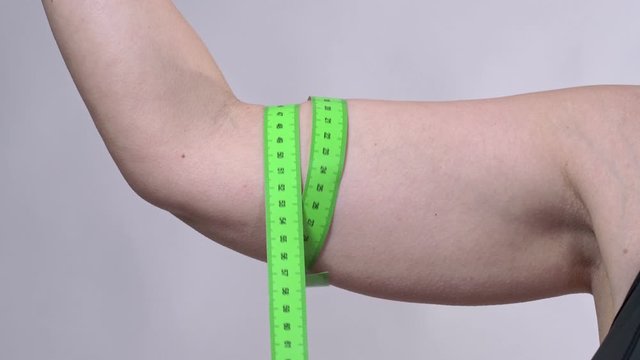 A middle-aged woman with saggy skin determines the circumference of her hand with a measuring tape, on a white background.