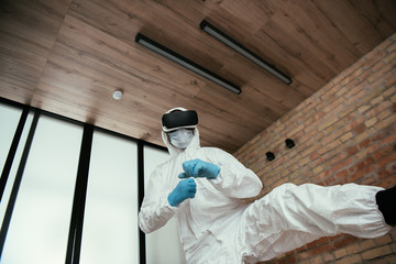 low angle view of man in hazmat suit, medical mask, latex gloves and virtual reality headset gaming in living room