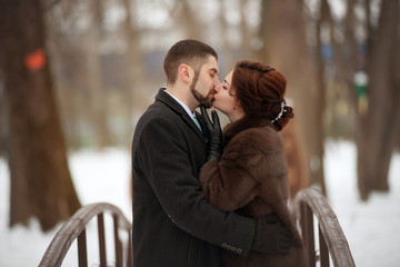 A bride in a fur coat kisses her bearded groom in a winter park.