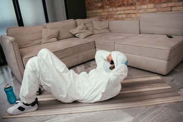 Fototapeta na wymiar man in hazmat suit, medical mask and goggles exercising near sports bottle in living room