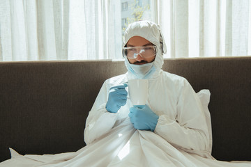 man in hazmat suit, headphones, medical mask, latex gloves and goggles holding cup in bed