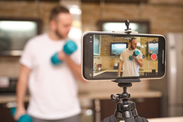 Athletic man blogger in sportswear shoots video on phone as she does exercises at home in the kitchen. Stay at home activities. Prevent infection from the pandemia.