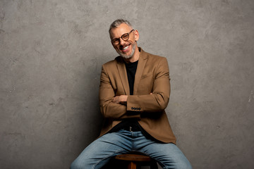 bearded businessman in glasses looking at camera and sitting with crossed arms on grey