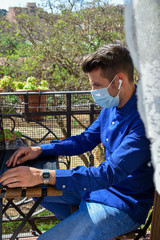 Obraz premium Man wearing a face mask making a video call with the laptop on his balcony, confined at home by the quarantine due to covid-19