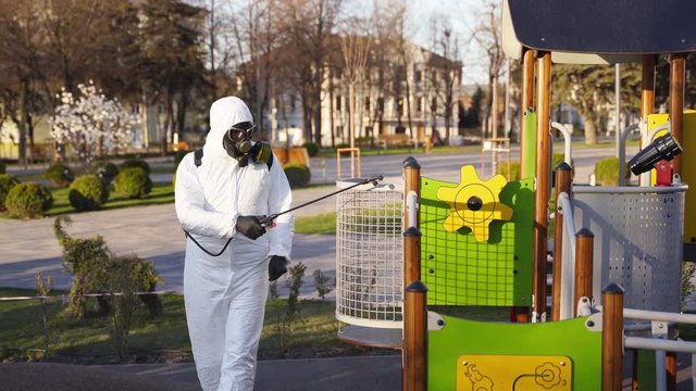 Hazmat team disinfects playground surfaces on coronavirus covid-19 quarantine with antibacterial sanitizer sprayer. Worker in gas mask and protective suit decontaminates children rides, slide, street.