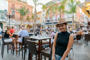 Happy young beautiful Asian tourist woman at the restaurant outdoors - obrazy, fototapety, plakaty