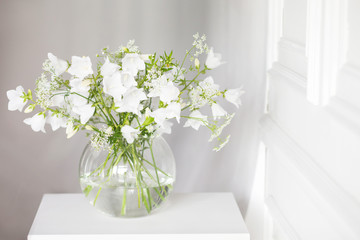 Bouquet of gentle bells in vase. Morning light in the room. Soft home decor, glass vase with white flowers on  white wall background and on wooden table. Interior. Greeting card. Copy space.