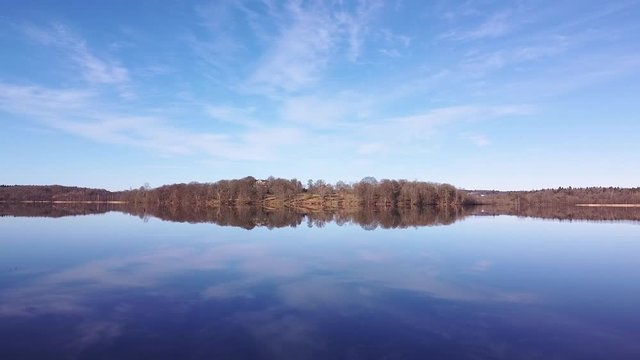 Slow aerial drone shot, flying from shoreside reeds towards a distant shore. A clear sky and distant shore are reflected in the mirror-like surface of a still lake at N&auml;&auml;s Fabriker, Tollered, Sweden.