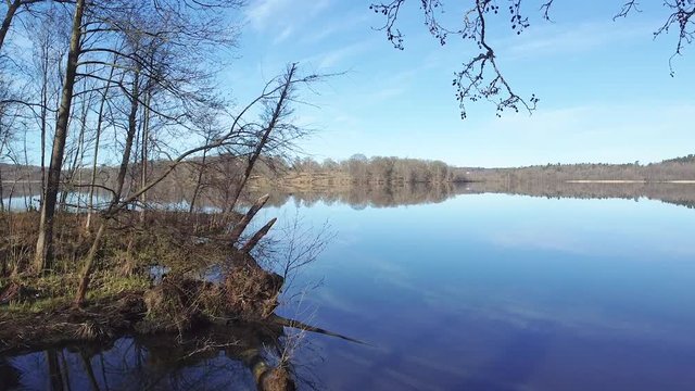 Slow aerial drone shot, flying by trees on the left shoreside, passing close to tree branches. A distant shore are reflected in the surface of a still lake at N&auml;&auml;s Fabriker, Tollered, Sweden.