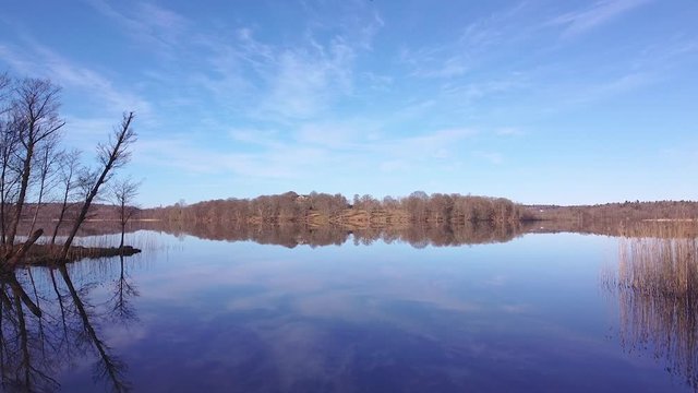 Slow aerial drone shot, flying by the shoreside, towards a distant shore. Clear sky, trees, reeds and a distant shore are reflected in the surface of a still lake at N&auml;&auml;s Fabriker, Tollered, Sweden.
