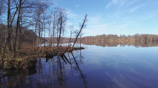 Slow aerial drone shot, flying beside early spring trees on the left shoreside. Sky, trees, reeds and a distant shore are reflected in the surface of a still lake at N&auml;&auml;s Fabriker, Tollered, Sweden.