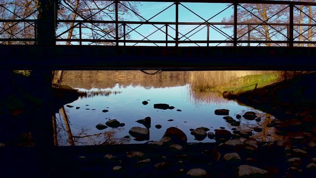 Slow aerial drone shot, flying close to a stony shore and under a bridge and out over the shoreline. A distant shore are reflected in the surface of a still lake at N&auml;&auml;s Fabriker, Tollered, Sweden.