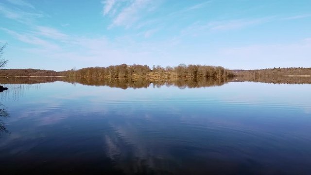 Slow aerial drone shot, flying from the shoreside towards a distant shore . A few white wispy clouds are reflected in the mirror-like surface of a still lake at N&auml;&auml;s Fabriker, Tollered, Sweden.