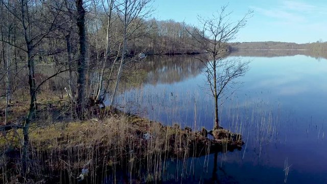 Slow aerial drone shot, flying by early spring trees on the shoreside, passing close by branches. A distant shore are reflected in the surface of a still lake at N&auml;&auml;s Fabriker, Tollered, Sweden.