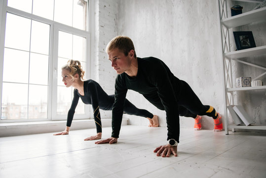 The guy and the girl in tracksuit. Black sports uniform. Male and female athletes. Pumped up body. Push ups. Morning work-out. Set of exercises for body. Classes in pair. Workout together at home.