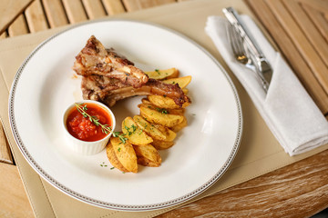 Close-up on a wooden table on a napkin is a large white round plate with dinner. For lunch, yellow Idaho country potatoes with grilled meat, ribs, red sauce, tomato ketchup
