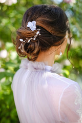 Naklejka premium Close-up hairstyle bun, collected dark hair, a young girl bride stands back in a lilac wedding dress against a background of green leaves. Beautiful white hair clip with a flower