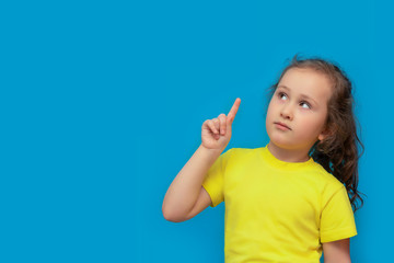 Emotional portrait of a preschool girl on a blue background. A little girl with long, curly hair points up with her index finger. Mock up. copy space