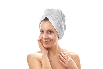 A beautiful woman with a white towel on her head does facial rejuvenation with eye patches and is smiling at the camera. White isolated background.