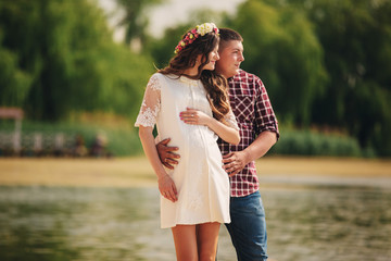 Young happy romantic pregnant couple hugging on nature near lake in summer park. Pregnant woman expecting a baby. Future mom and dad, family. mother's, father's day