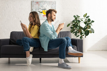 Fototapeta premium Side view of couple using smartphones while sitting back to back on sofa