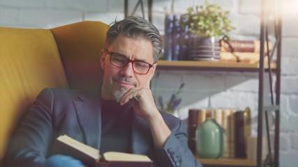 Older white man in glasses reading book at home sitting in armchair in cosy room.