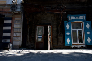 The old house of timber with open blue shutters and an open wooden door stands on the city street. Irkutsk
