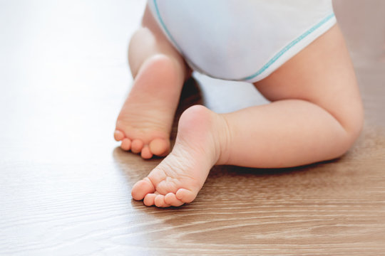 Baby's feet on wooden floor. Bare heels of little child wearing white bodysuit and diaper. Cozy morning bedtime at home.