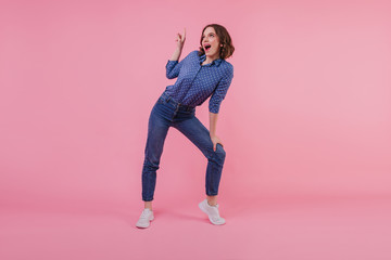 Full-length portrait of pleased white girl in trendy denim pants. Indoor photo of smiling good-humoured woman posing on pink background.