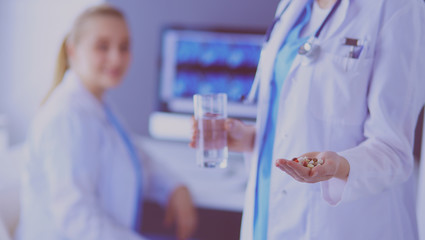 shot of doctor holding pills and glass of water at clinic with colleague on the background.