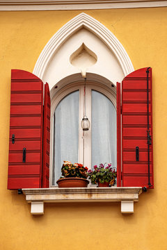 Close-up of a window with gothic arch and open red shutters on an orange wall. Caorle, Venice province, Veneto, Italy, Europe