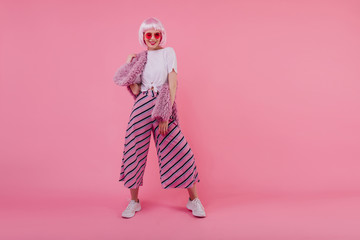 Full-length portrait of confident young woman in trendy pants. Indoor shot of inspired lady in peruke and sunglasses isolated on bright pink background.