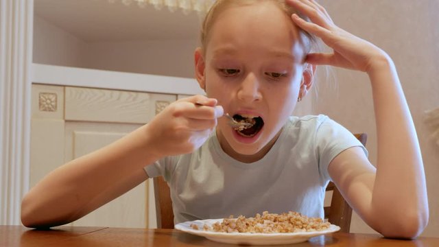 Sad girl reluctantly eating porridge at kitchen. Low angle view of upset young girl holding hand on head while eating unloved food. Food concept