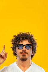 Portrait of happy bearded man with curly hair pointing finger up at copyspace isolated over yellow background. Guy in blue sunglasses pick on great idea.