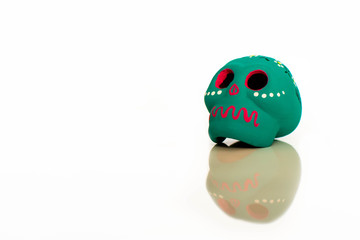A Mexican day of the dead mask or small model.  The icon sits on top of a reflective surface creating a mirror image.  The bright green mask decorated in vivid colours, sits against a plain background