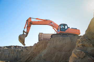  orange crawler excavator and a construction dump truck at work on a sand quarry, standing next to it while working on a sunny day against a blue sky. View from below