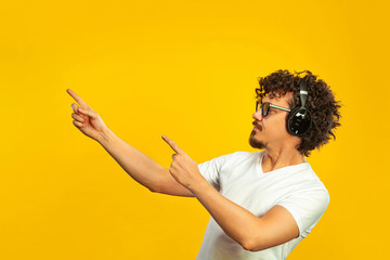 Portrait of happy bearded man with curly hair pointing finger at screen isolated over yellow background. Guy in blue sunglasses and headphones pick on great idea.