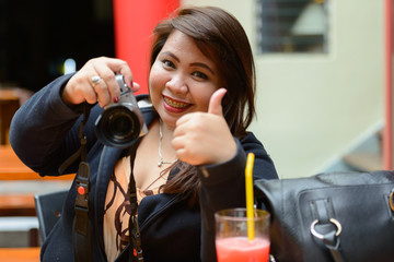 Happy young overweight Asian woman taking picture and giving thumbs up