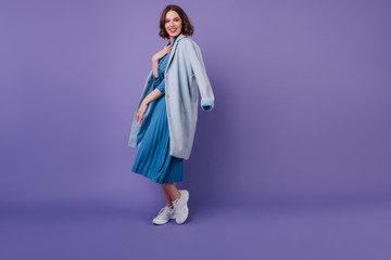 Full-length shot of fashionable girl in white sneakers wears blue coat. Indoor portrait of carefree...