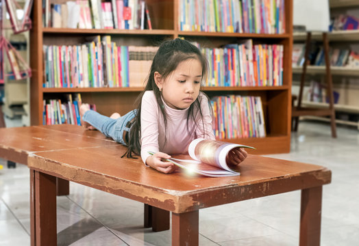 Little girl laying down on wooden table,reading book ,with interested feeling,at school,blurry light around,Leens flare effect