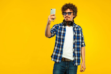 European man with curly hair in blue sunglasses with mobile phone or smarphone. Handsome smiled stylish hipster in plaid shirt posing over yellow background.