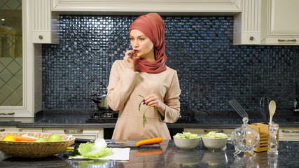 charming oriental woman in hijab and long dress stands at kitchen table and eats greens preparing food for family closeup