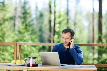 Serious middle aged man talking on cell phone and using laptop computer sitting at wooden table on country house balcony. Man working remotely online