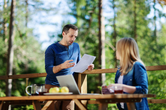 Middle Aged Couple Planning Home Finances Or Discussing Bill On Country House Balcony. Man Reading Document To Wife Sitting At Wooden Table With Open Laptop