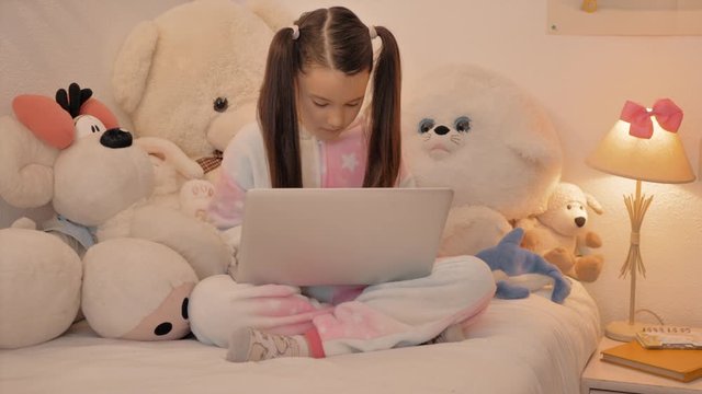 ni&ntilde;a con coletas en pijama rosa sentada en su cama rodeada de peluche estudia con un port&aacute;til