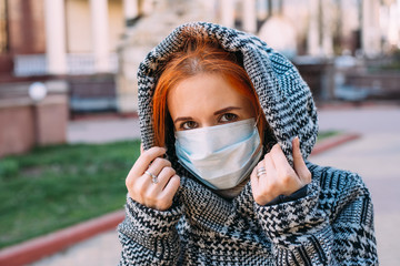 A young red-haired woman wearing protective medical mask and hood of a gray coat during coronavirus pandemic - 342417093