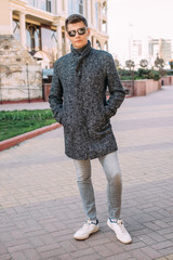 A handsome young man in a stylish grey coat, grey jeans and sunglasses stands on the street - 342417441